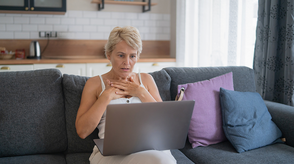 A woman looks shocked and concerned, placing her hands on her chest while staring at her laptop screen in a cozy living room.